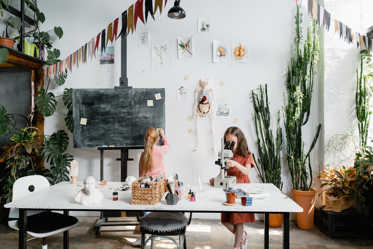 Two young girls explore science with a microscope and skeleton model in a classroom filled with plants.