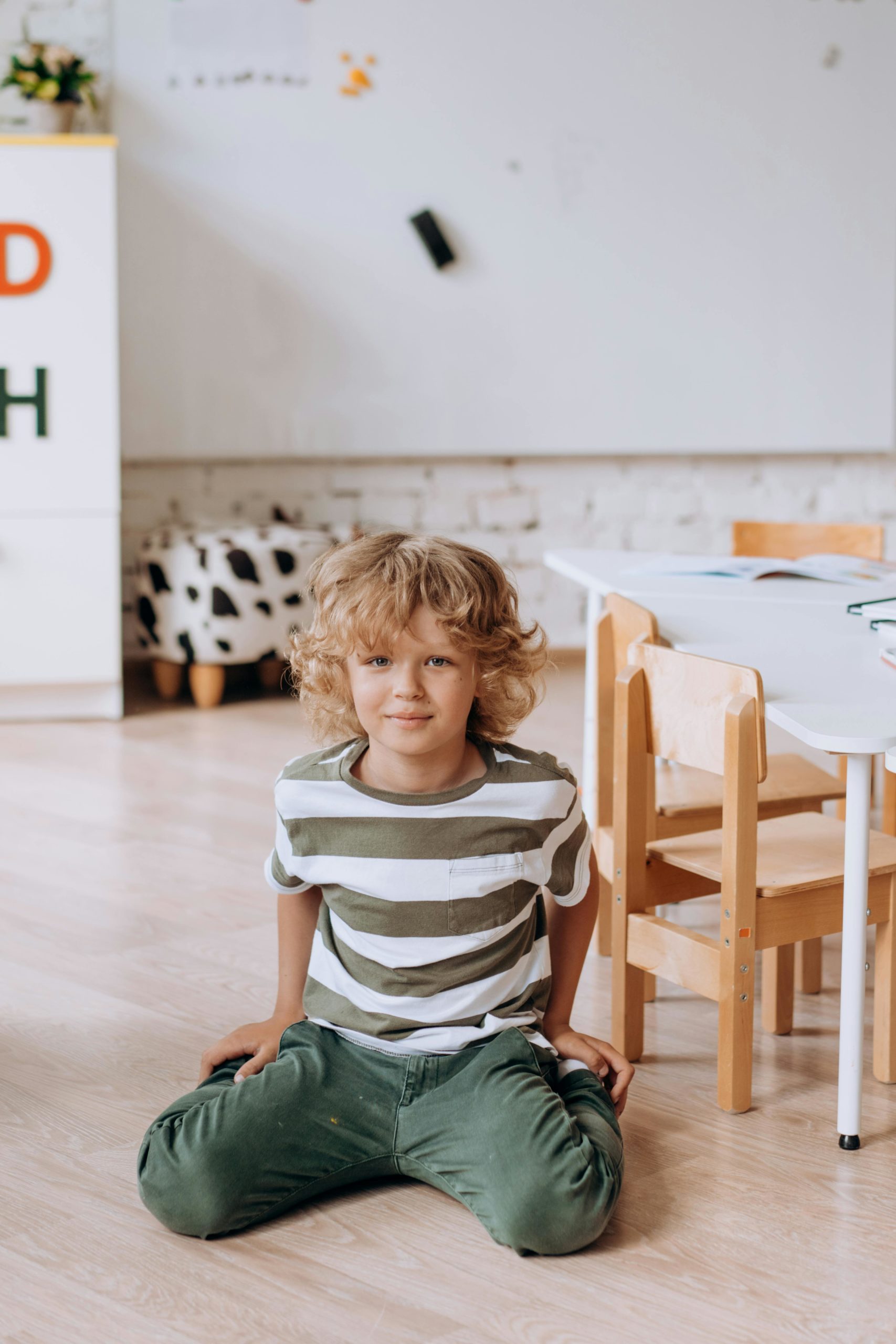 A cheerful young boy with blond hair sitting on the floor in a classroom, wearing a striped shirt.